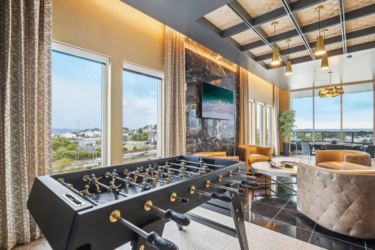 An airy resident lounge with a foosball table, plush seating, expansive windows offering dramatic views, and colorful wall art. The atmosphere is playful yet sophisticated.
