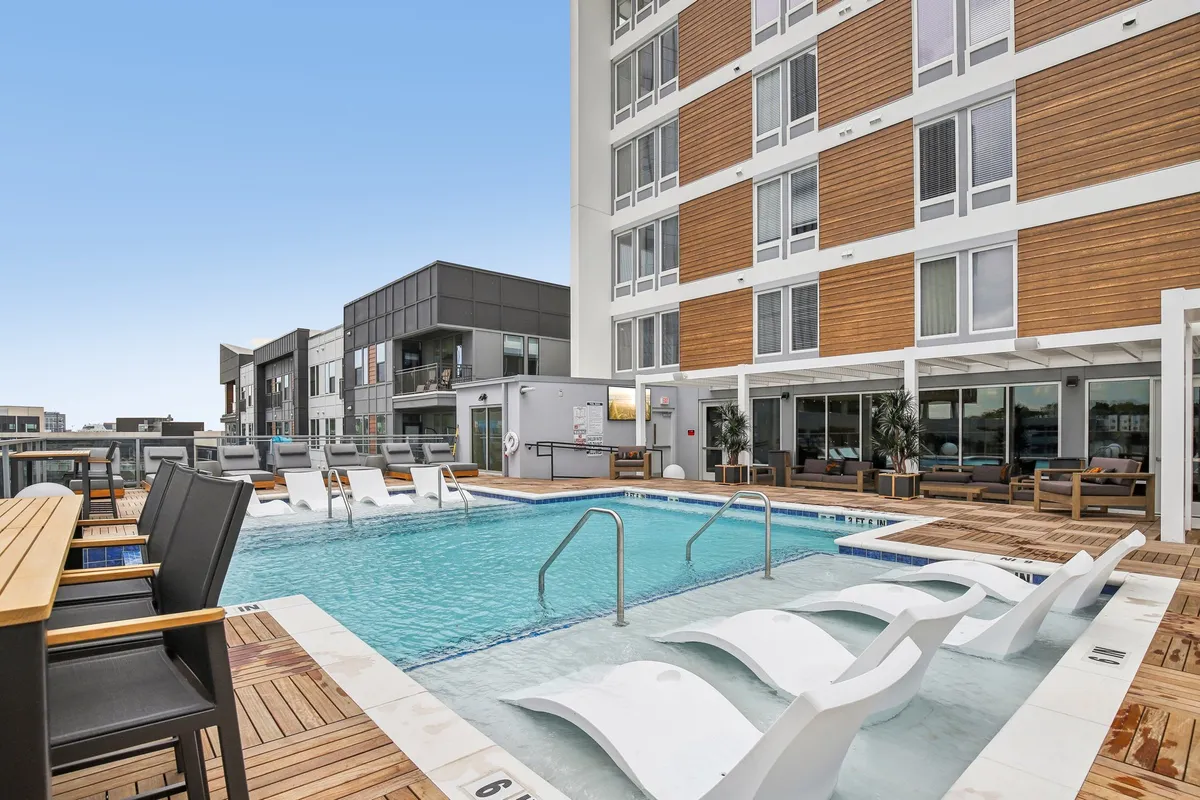 A sunlit swimming pool surrounded by wooden decking, modern lounge chairs, and urban buildings in the background. The scene captures people relaxing poolside and enjoying the open-air ambiance.