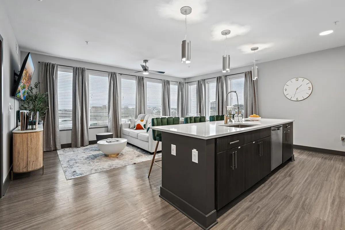 A contemporary, open-concept living space featuring a welcoming kitchen island with green chairs, a cozy sofa, large windows that let in natural light, and a decorative rug. The layout is perfect for family gatherings, meals, and casual relaxation.