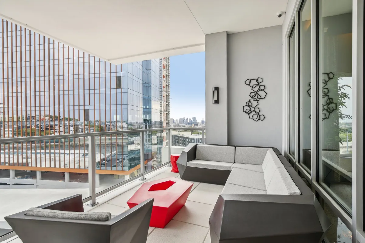 A stylish outdoor lounge area featuring modern seating, a bold red geometric coffee table, and sweeping views of the city skyline. Decorative wall art adds a contemporary touch.