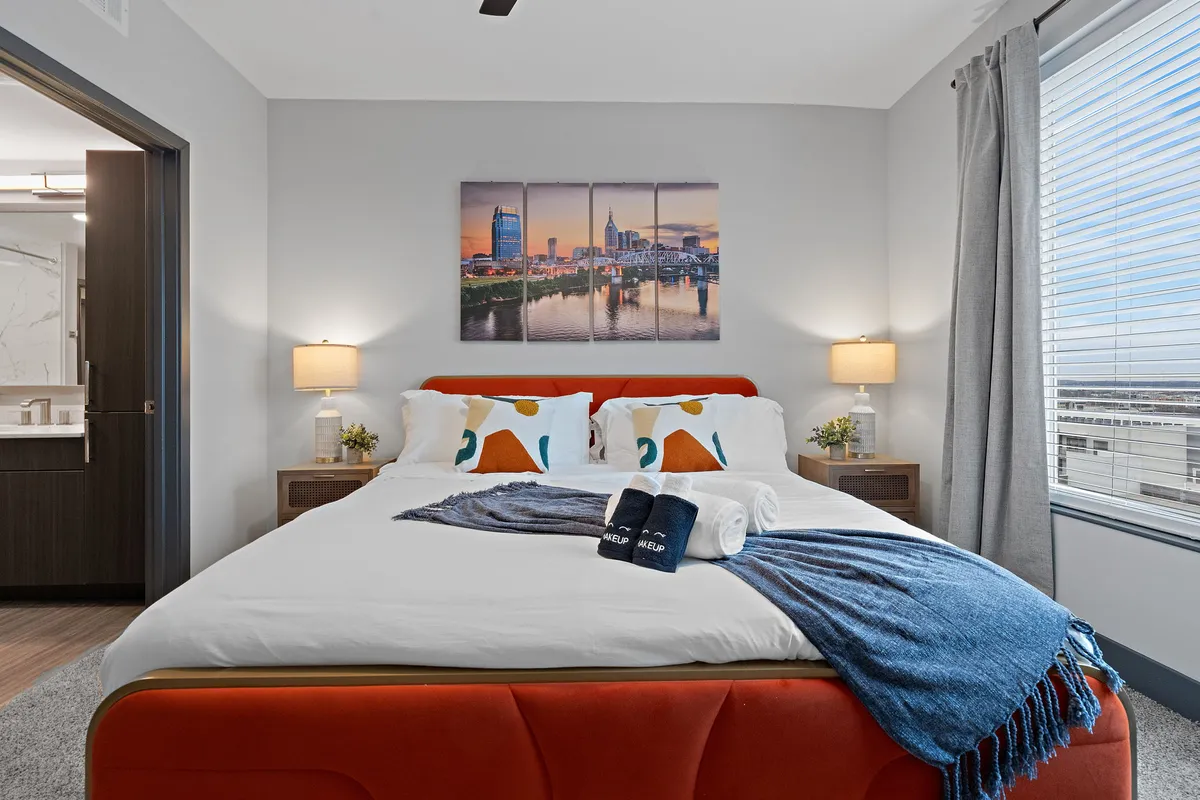 A stylish bedroom featuring a plush bed adorned with colorful pillows and a thick blue throw blanket. Large artwork of a cityscape hangs above the bed, while nightstand lamps provide gentle lighting. The ambiance is comfortable and inviting, ideal for relaxing or waking up to sunlight through the window.