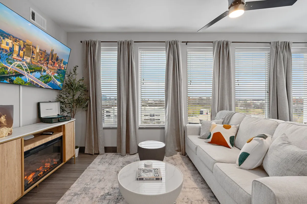 A stylish living room featuring a large TV displaying a vibrant cityscape, a modern sofa adorned with decorative pillows, and a sleek fireplace. The arrangement suggests a cozy spot to unwind while enjoying sweeping views from the window, with plenty of natural light setting a romantic ambiance.