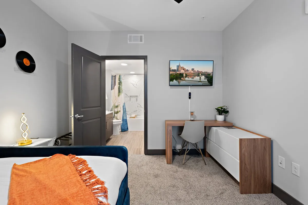 A modern, comfortable room with a bed adorned by a vibrant orange throw, a workstation with a chair, a TV mounted on the wall, and a view towards the bathroom. The setup is ideal for both productivity and relaxation, offering a dedicated space for working on projects or winding down.