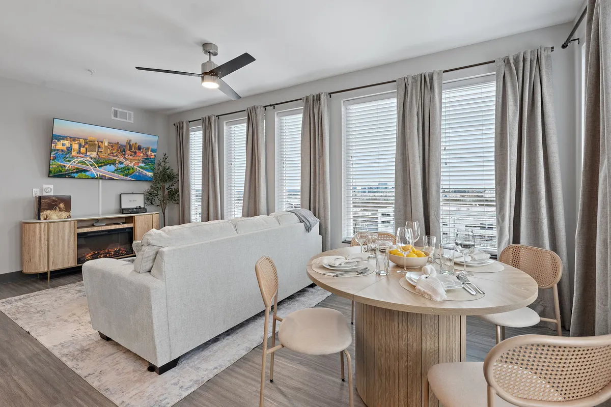 An inviting open-concept living and dining space with a plush sofa, a modern dining table set for four, a large TV, and expansive windows that allow natural daylight to stream in. The layout clearly showcases both the living and dining areas as one cohesive space.