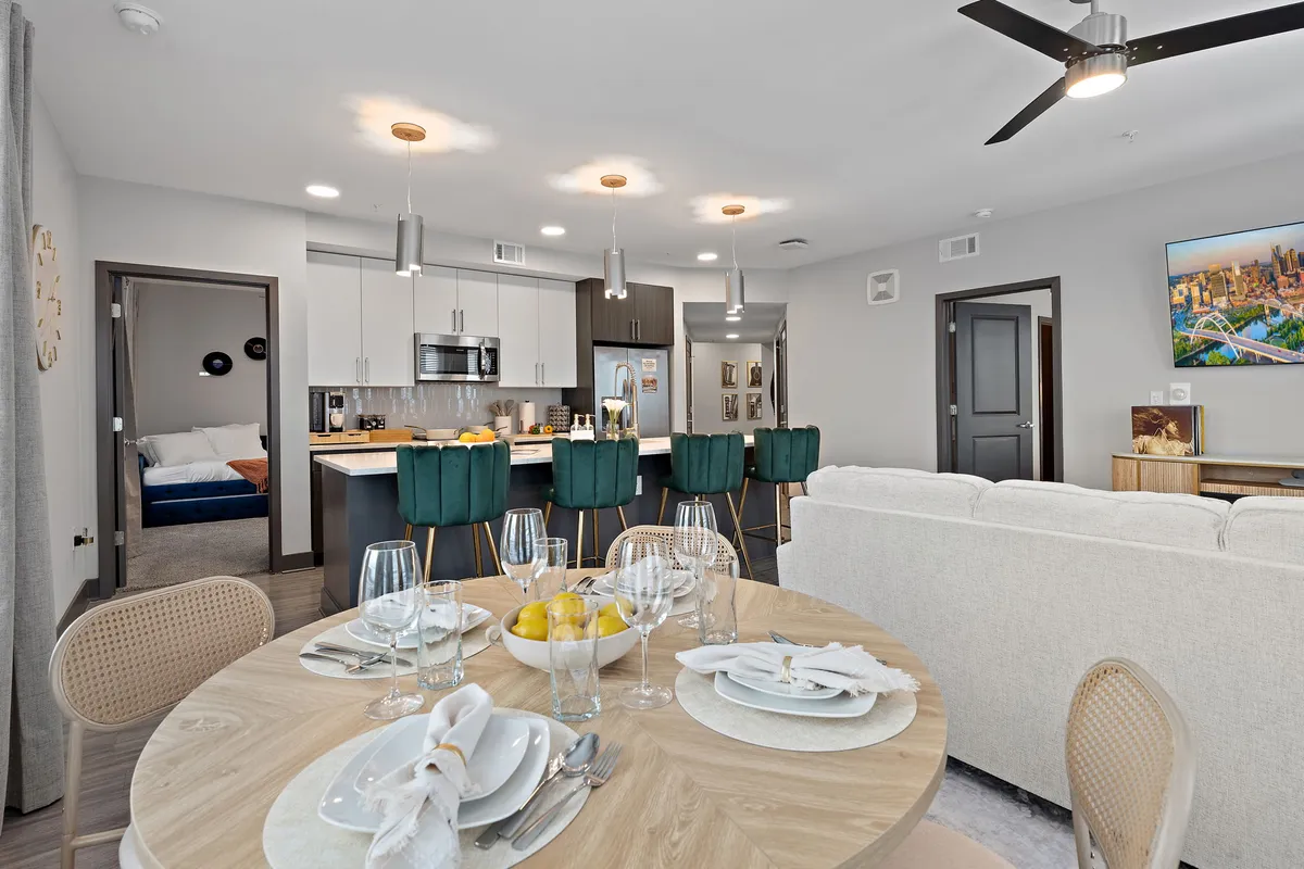 A broader perspective of the living space, featuring a dining table set with place settings, a kitchen island with bar stools, a comfortable sofa, and wall art. The design is open and contemporary, with natural light filling the space.