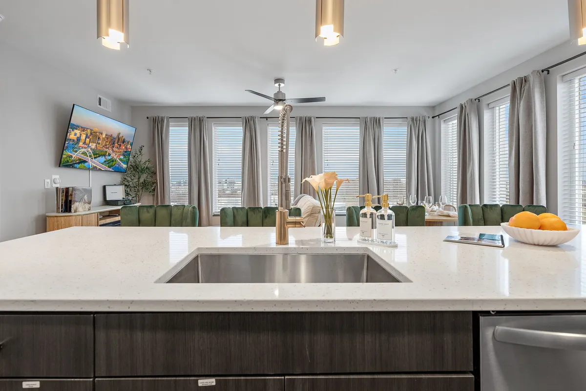 A modern, open-concept kitchen and living area with floor-to-ceiling windows that frame a panoramic view of the city. The space includes a stylish kitchen island with green chairs, a cozy seating area, and a television displaying a city scene, reinforcing the urban vibe.