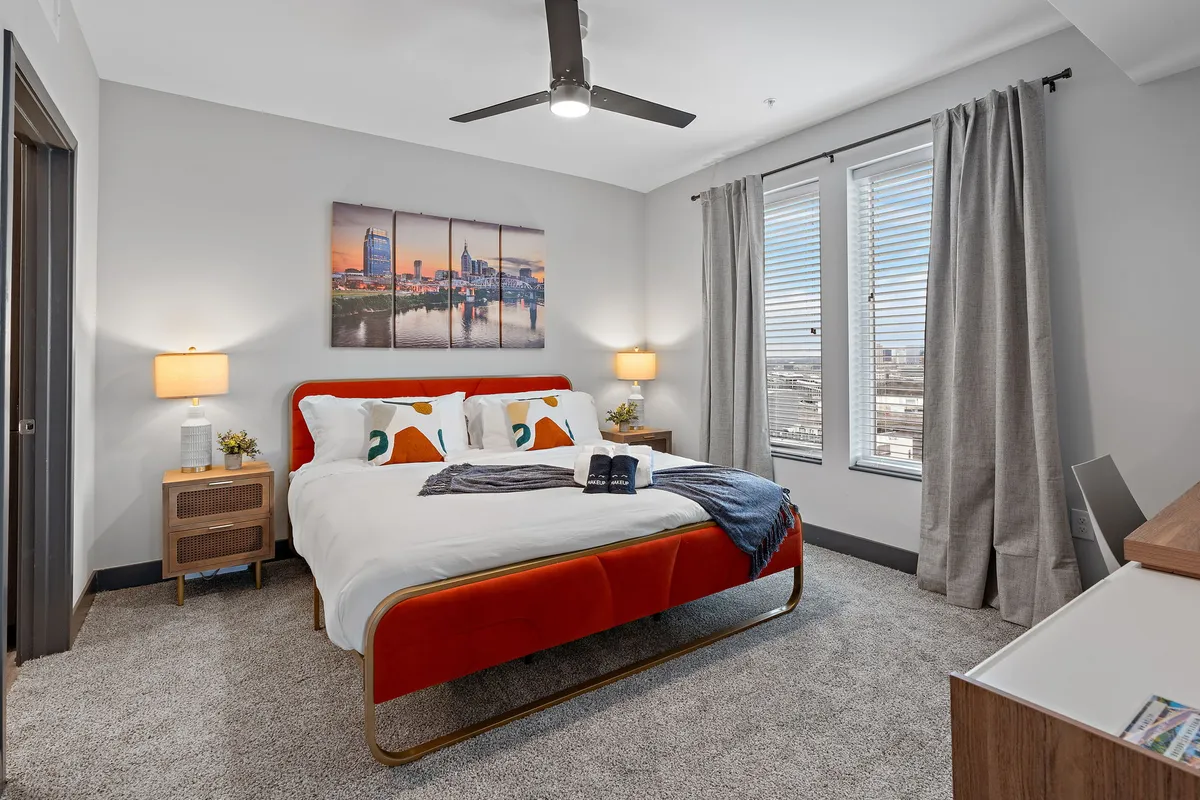 The image showcases a stylish bedroom with a plush bed adorned with vibrant, colorful pillows. Modern nightstands with lamps flank the bed, and eye-catching artwork brings energy to the space. Morning sunlight streams through the window, illuminating the cozy, contemporary decor and creating an inviting, restful atmosphere.