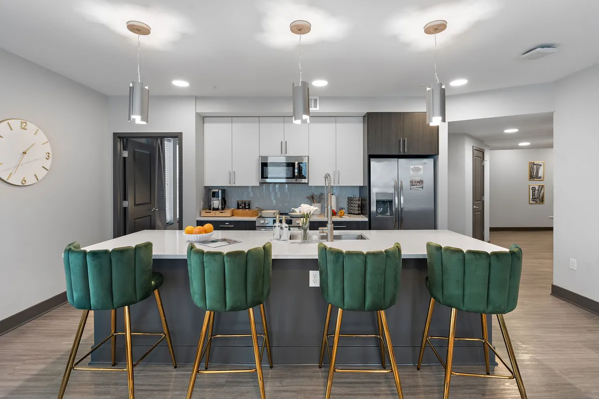 A bright and modern kitchen scene with green bar stools lining a marble countertop, complemented by modern kitchen appliances and a prominent wall clock. The space feels welcoming and stylish, suggesting easy breakfasts or casual meals.