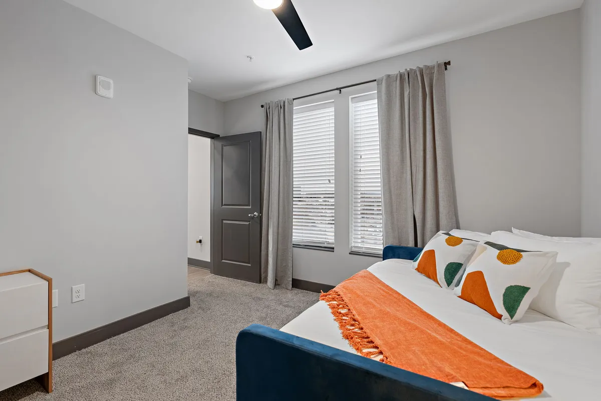 A cozy bedroom highlighted by a bed with colorful pillows and an orange throw blanket, set beside large windows with curtains drawn to let in soft, natural light. The space feels tranquil and restful, with a focus on comfort and relaxation.